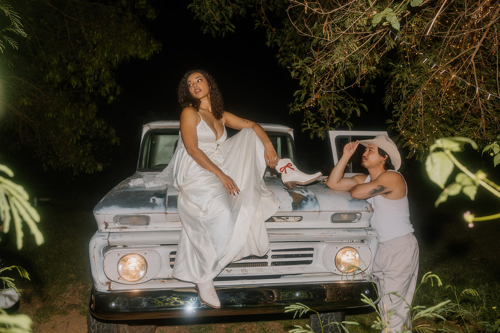 A Western bride in a flowing gown and white boots sits confidently on the hood of a vintage Chevrolet truck, while her groom, in a cowboy hat, leans on the hood looking up at her under a moody night sky.