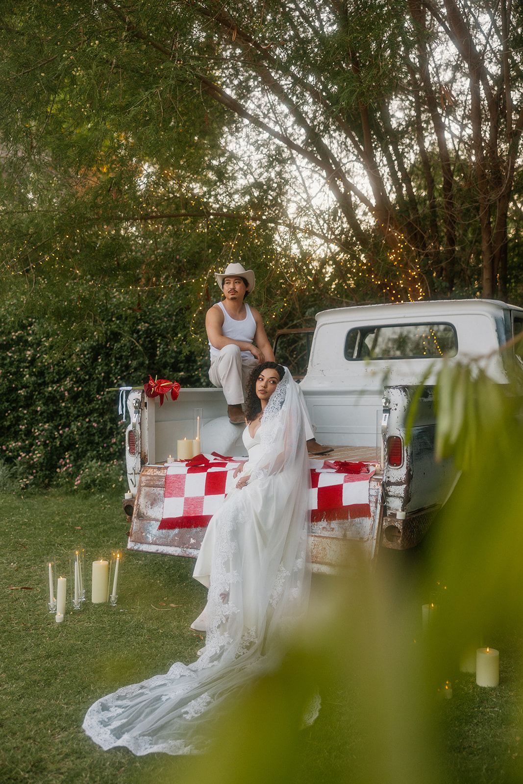 A romantic outdoor scene shows a groom sitting on the tailgate of a vintage white truck while the bride in a long white veil and dress sits below him, surrounded by candles and string lights. The truck bed is decorated with a red and white checkered blanket.