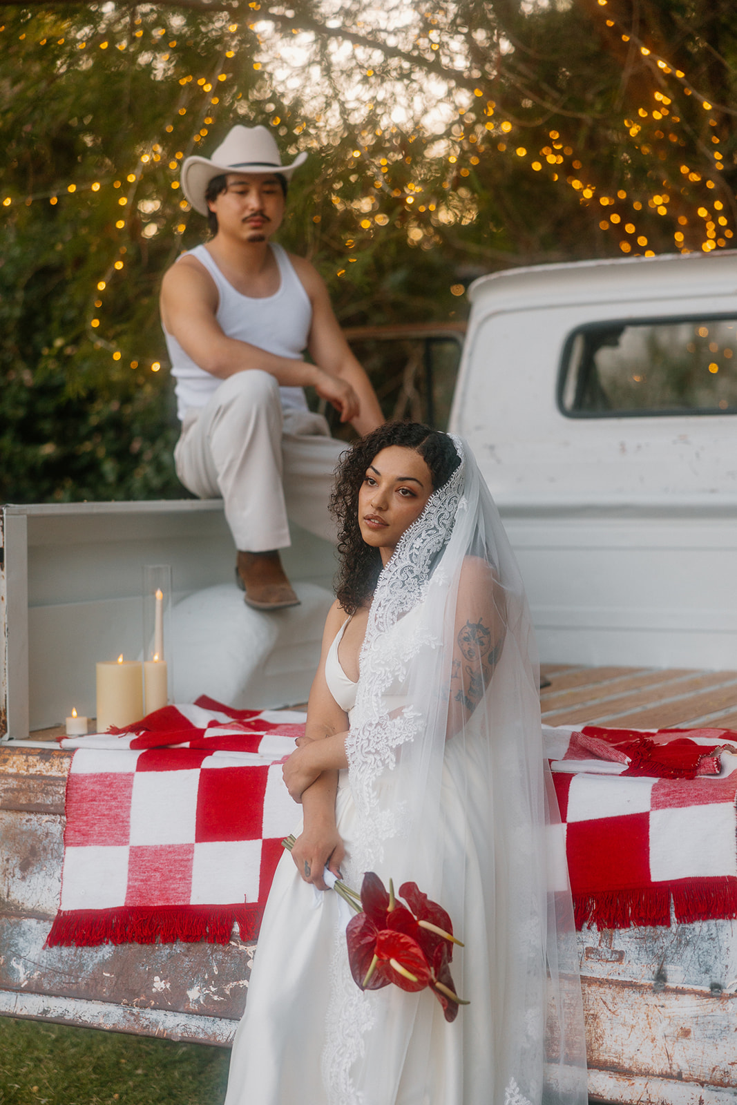 A bride with a cathedral-length veil and a moody bouquet of red anthuriums leans against an old pickup truck while her groom in Western attire perches above, both bathed in golden light and surrounded by string lights.