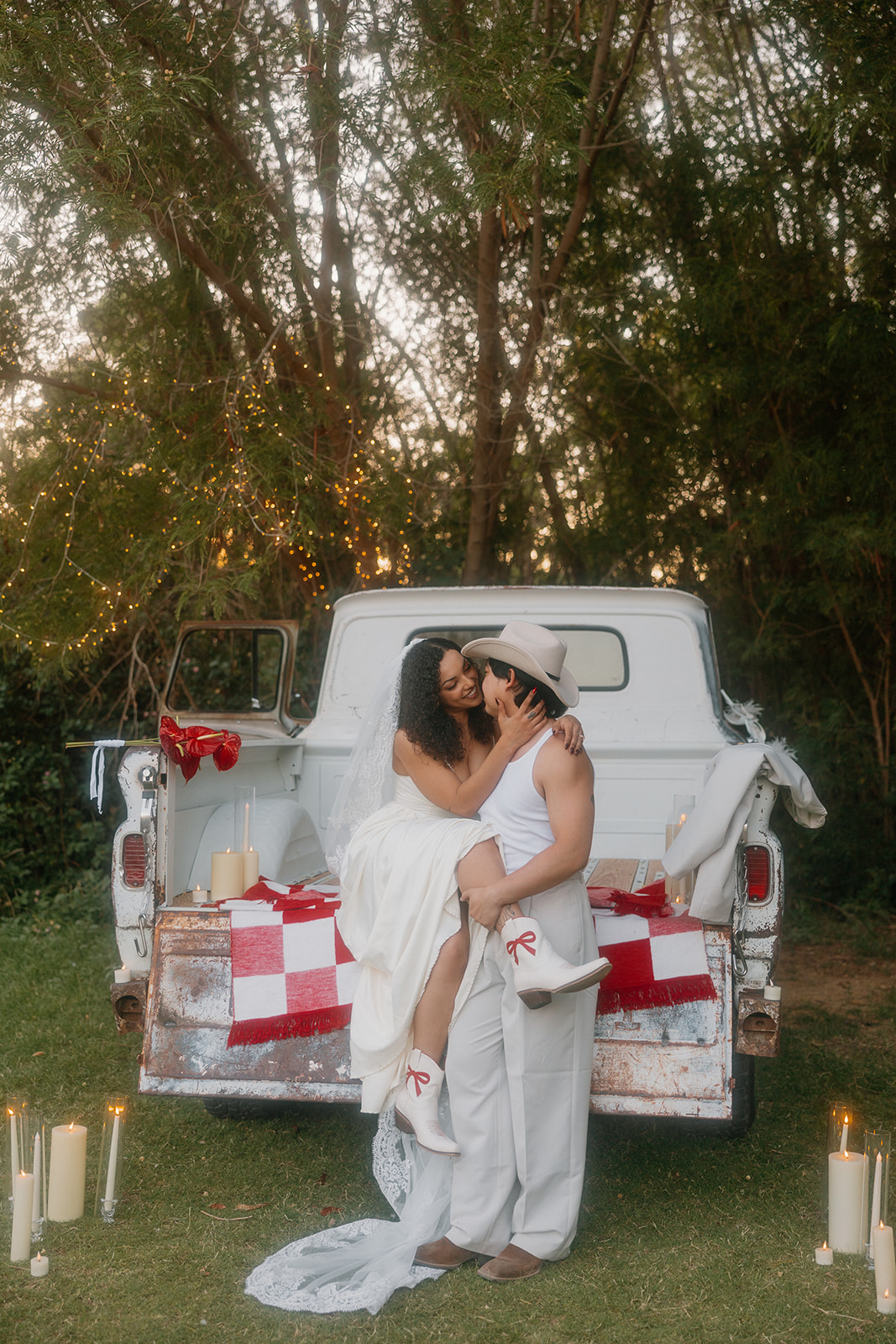 Bride and groom in a western wedding setting, sitting in an old white pickup truck with the bride in a white gown holding red flowers.