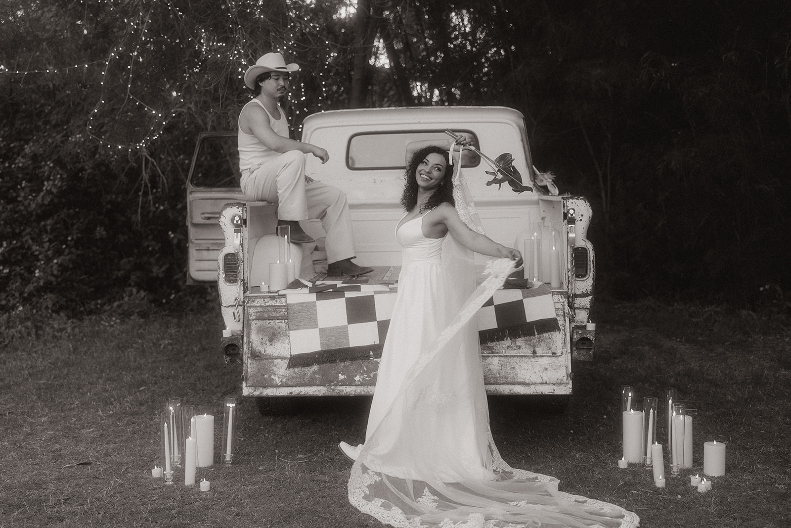 A black-and-white shot of a Western-inspired wedding scene featuring a joyful bride in a flowy gown and long veil, dancing in front of a vintage pickup truck adorned with candles and checkered textiles. A cowboy-hatted groom sits casually on the tailgate under fairy lights.
