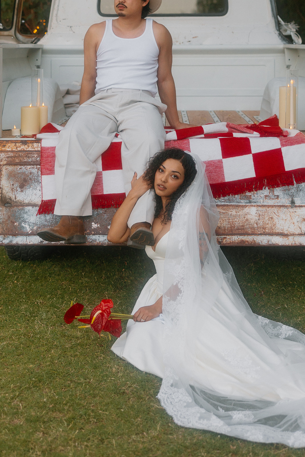 The bride sits on the grass in front of the truck, holding red flowers and looking at the camera. The groom sits above her on the tailgate, his leg draped beside her shoulder. The truck bed features a red and white picnic blanket and candles.