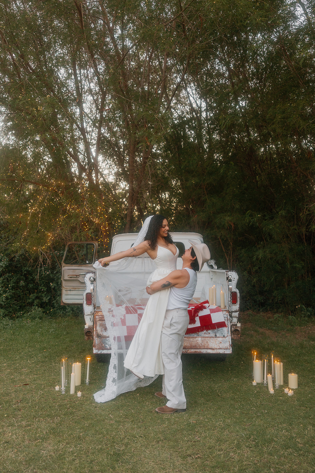 The groom lifts the bride in front of a vintage pickup truck, her veil floating midair as they smile at each other under string lights and surrounded by tall trees and candlelight.