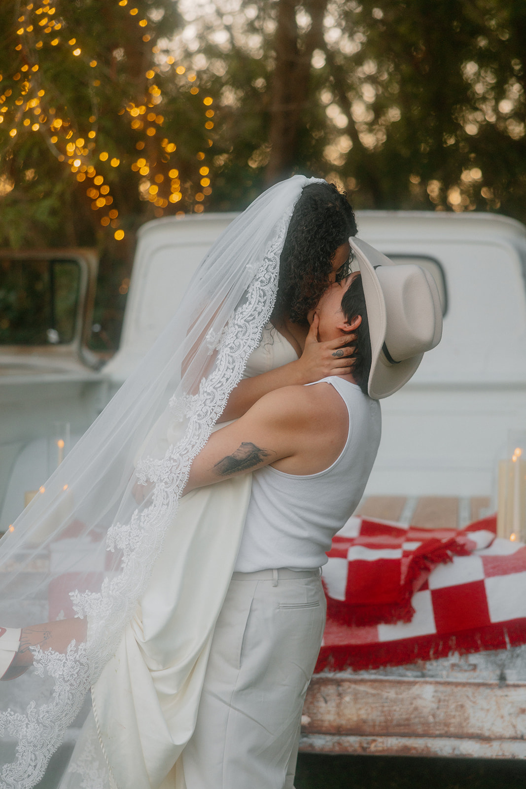 A groom in a cowboy hat lifts his bride for a kiss, her lace-trimmed veil cascading over both of them as they embrace in front of a vintage pickup bed draped with a red-and-white checkered blanket and surrounded by glowing candles.