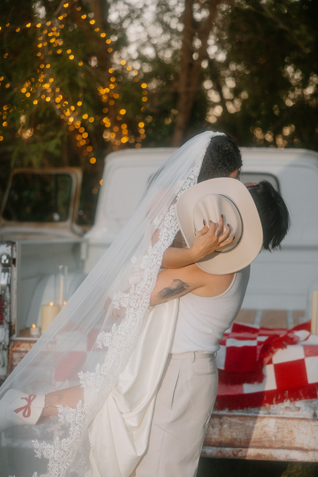 A close-up of a bride and groom in a passionate kiss, her fingers clutching his cowboy hat as she wraps her veil around them. The rustic truck in the background sets the perfect Western wedding scene.