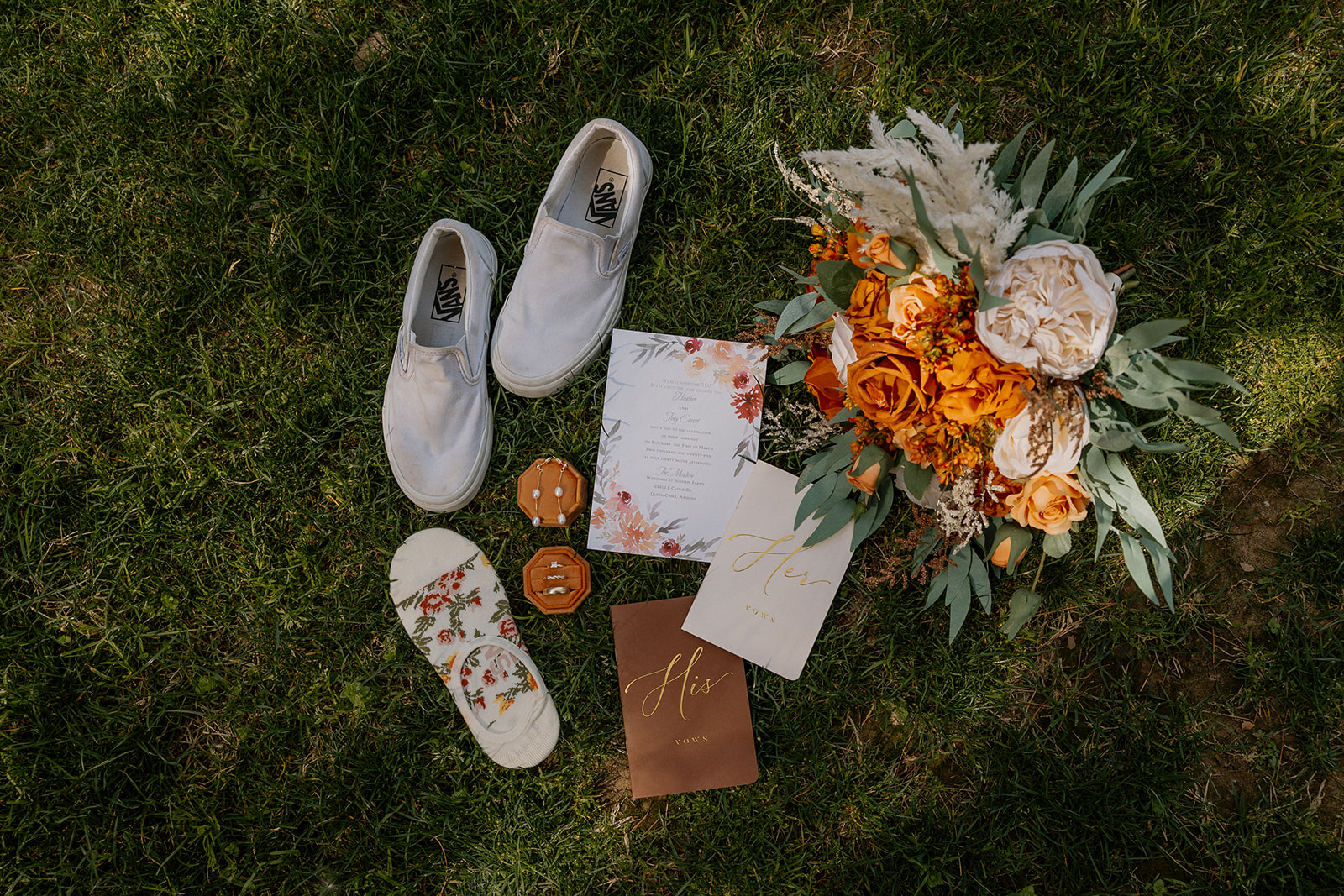 Flat lay of wedding day details including vow books, rings, floral socks, shoes, and a bouquet on the grass at Schnepf Farms.