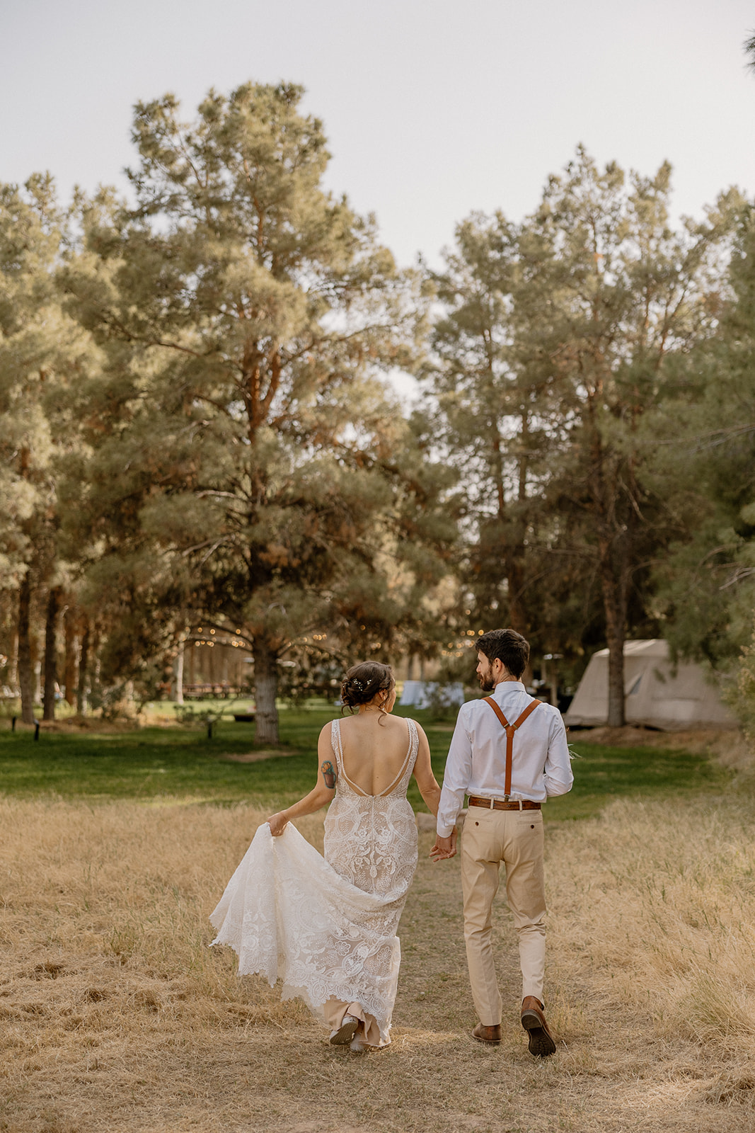 Bride and groom walk hand in hand through the golden grass at Schnepf Farms, trailing her lace dress behind.