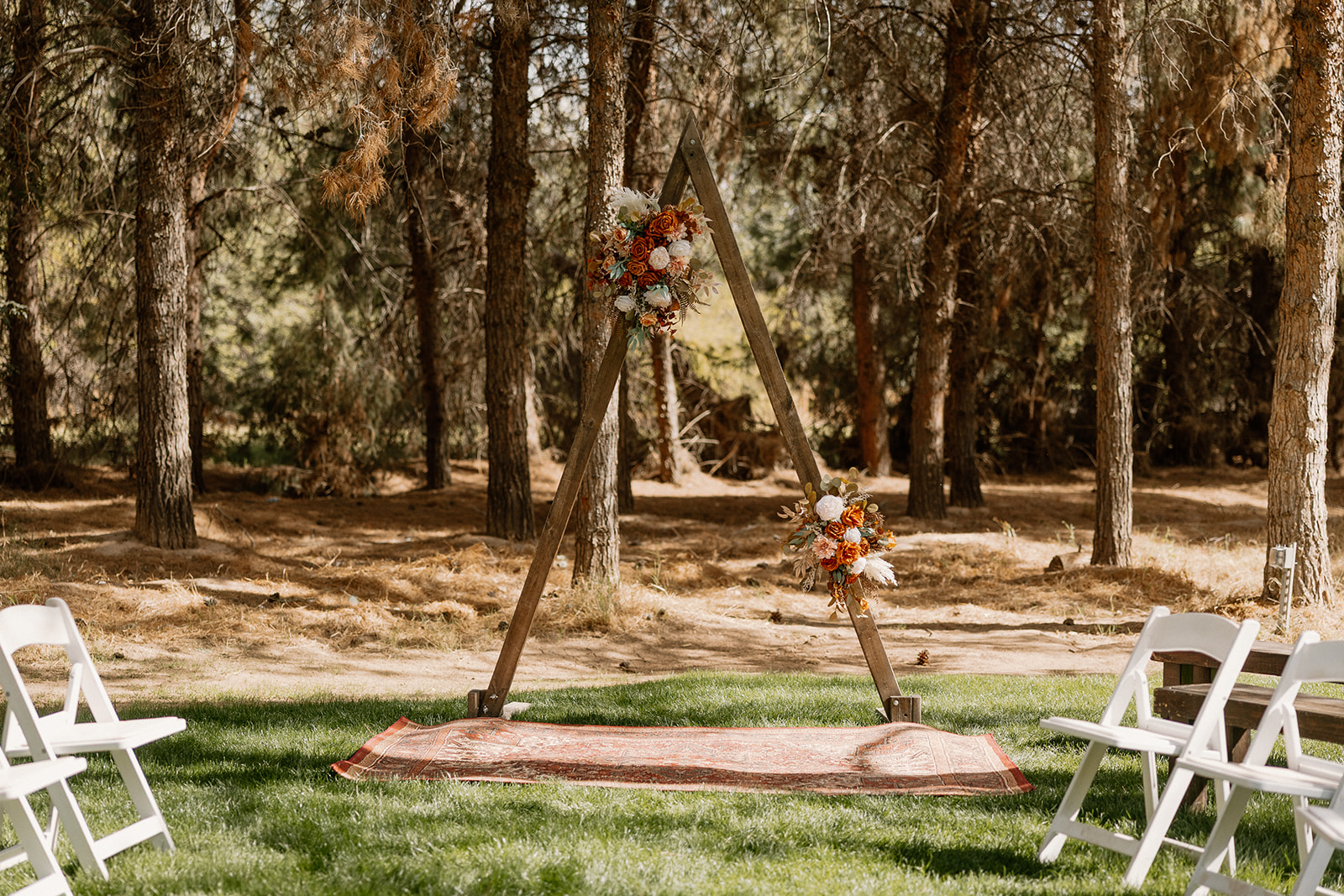 Wooden triangle wedding arch adorned with rust and cream florals set up in a shaded pine grove ceremony space at Schnepf Farms.