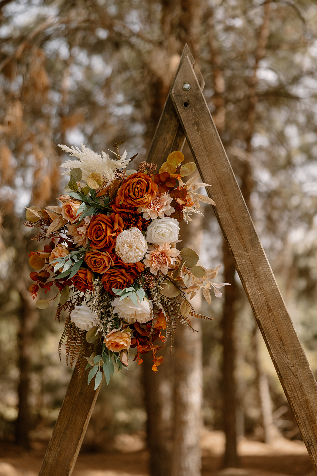Close-up of a fall-toned floral arrangement featuring roses, eucalyptus, and dried accents on a wooden triangle arch at Schnepf Farms.