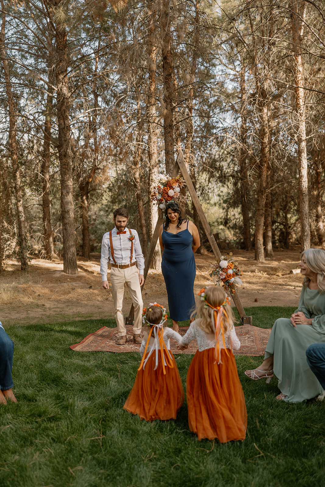 Flower girls in rust dresses walk toward the groom and officiant at the wooden triangle arch altar during a ceremony at Schnepf Farms.