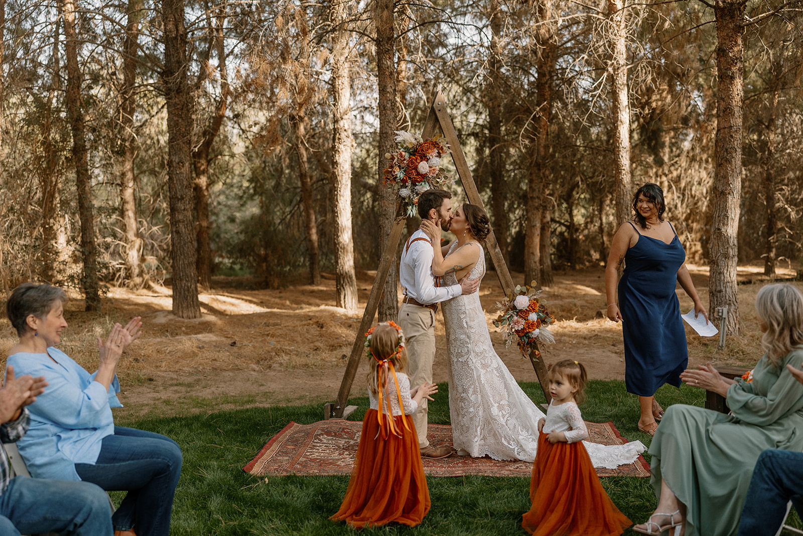 Bride and groom share their first kiss under a triangle arch while guests clap and flower girls wander in the foreground at Schnepf Farms.