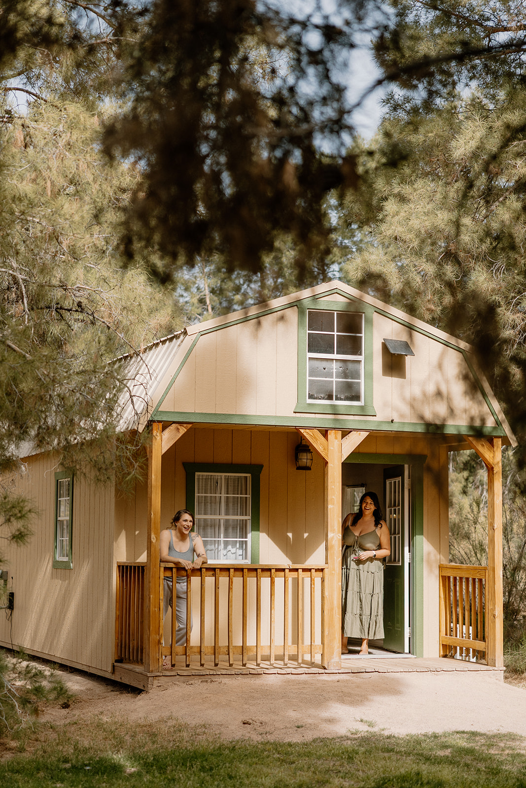 Two women relax on the front porch of a charming tan and green cabin tucked in the trees at Schnepf Farms.