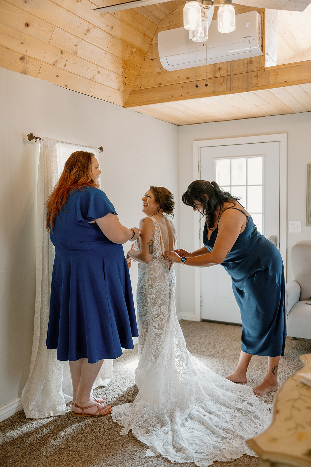 Bride laughing with her bridesmaids while getting buttoned into her lace wedding dress inside a cozy prep room at Schnepf Farms.