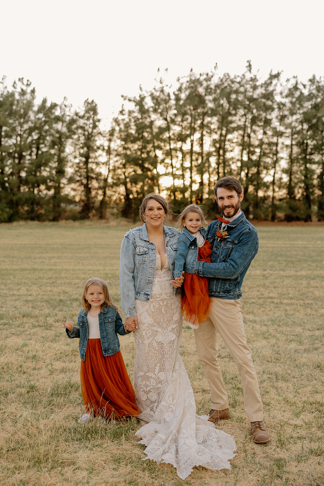 Bride and groom pose with their two daughters in matching denim jackets and rust dresses on a grassy field at sunset.