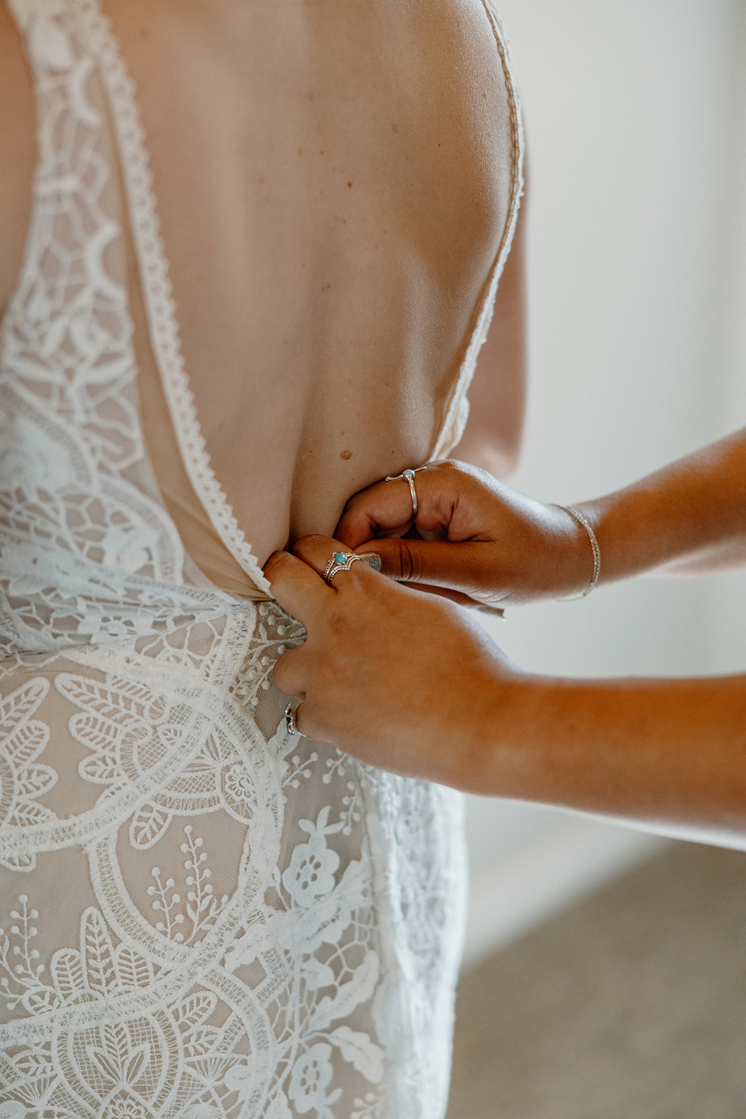 Detailed close-up of hands fastening the lace back of a bride’s wedding gown as she gets ready.