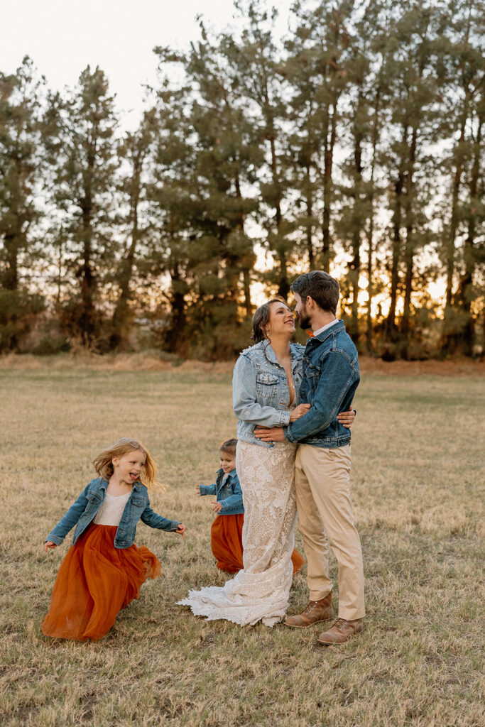 Bride and groom embrace while daughters run around them in matching flower girl orange dresses.