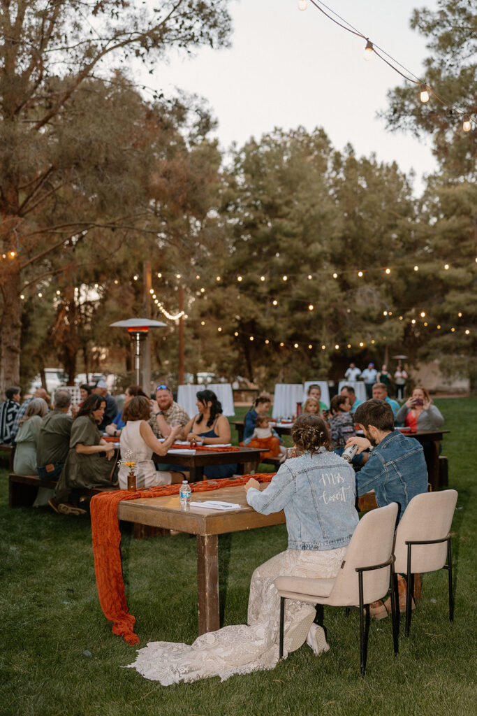 Bride and groom sit at sweetheart table surrounded by guests enjoying their dinner while celebrating the newlyweds. 