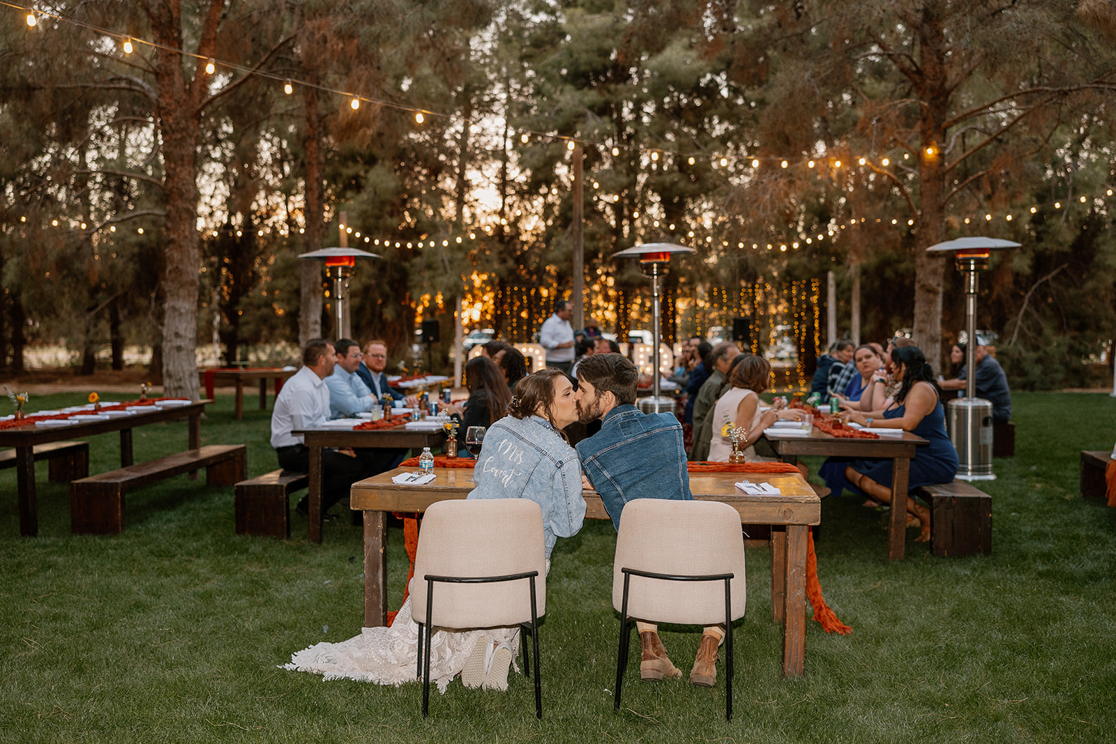 Newly married couple wearing jean jackets over wedding attire kiss at table during evening reception at Schnepf Farms