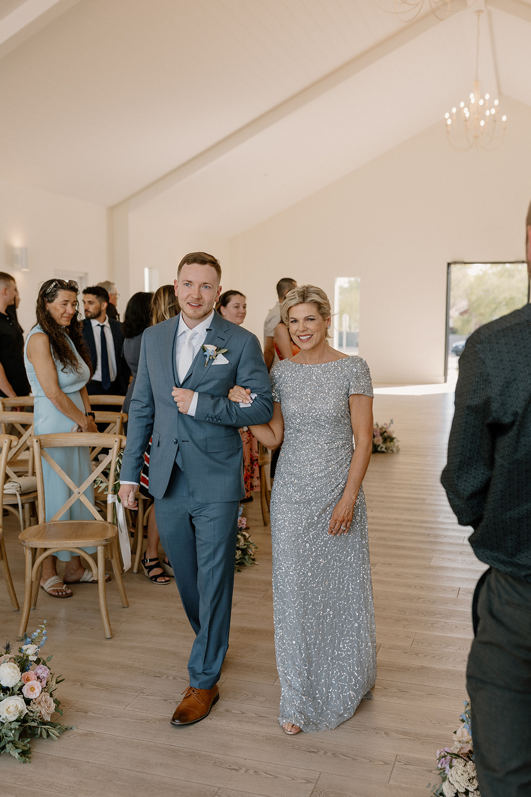 Groom walks down aisle with mother as guests stand to welcome him to the ceremony. 