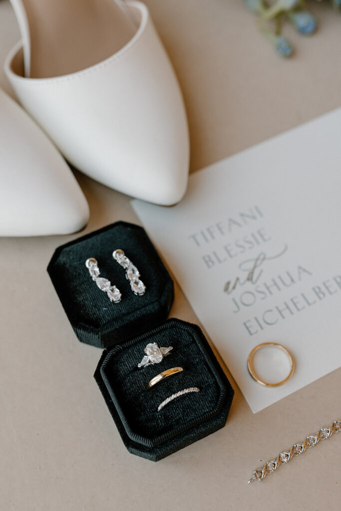 Close up of engagement ring and wedding bands sitting on invitations with bride's earrings and shoes behind them