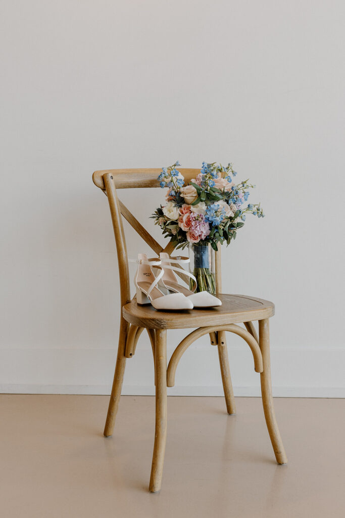 Bridal shoes sitting on chair next to bridal bouquet with pink, blue, and yellow florals throughout. 