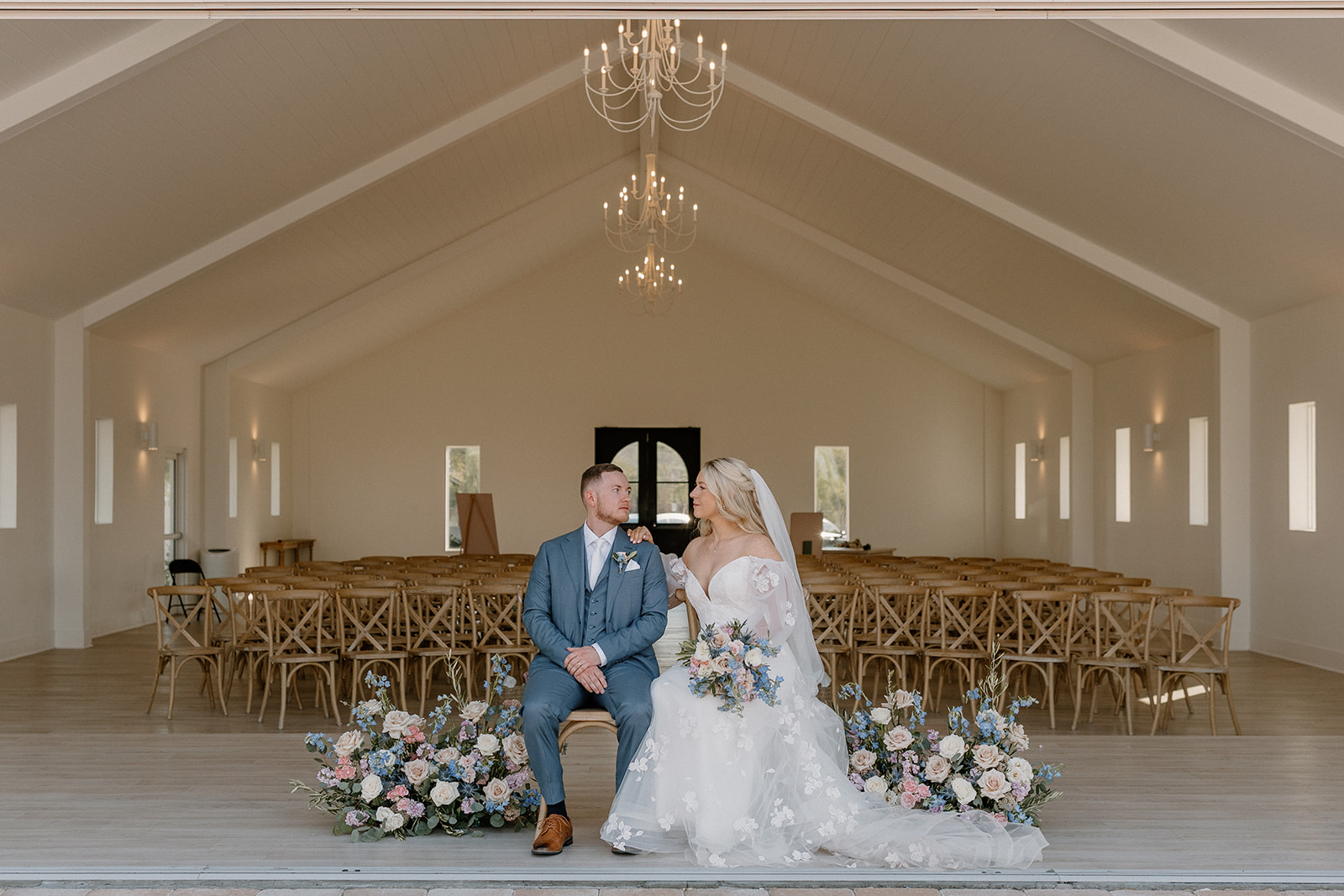 Wide shot of ceremony space with bride and groom sitting in chairs looking at one another. 