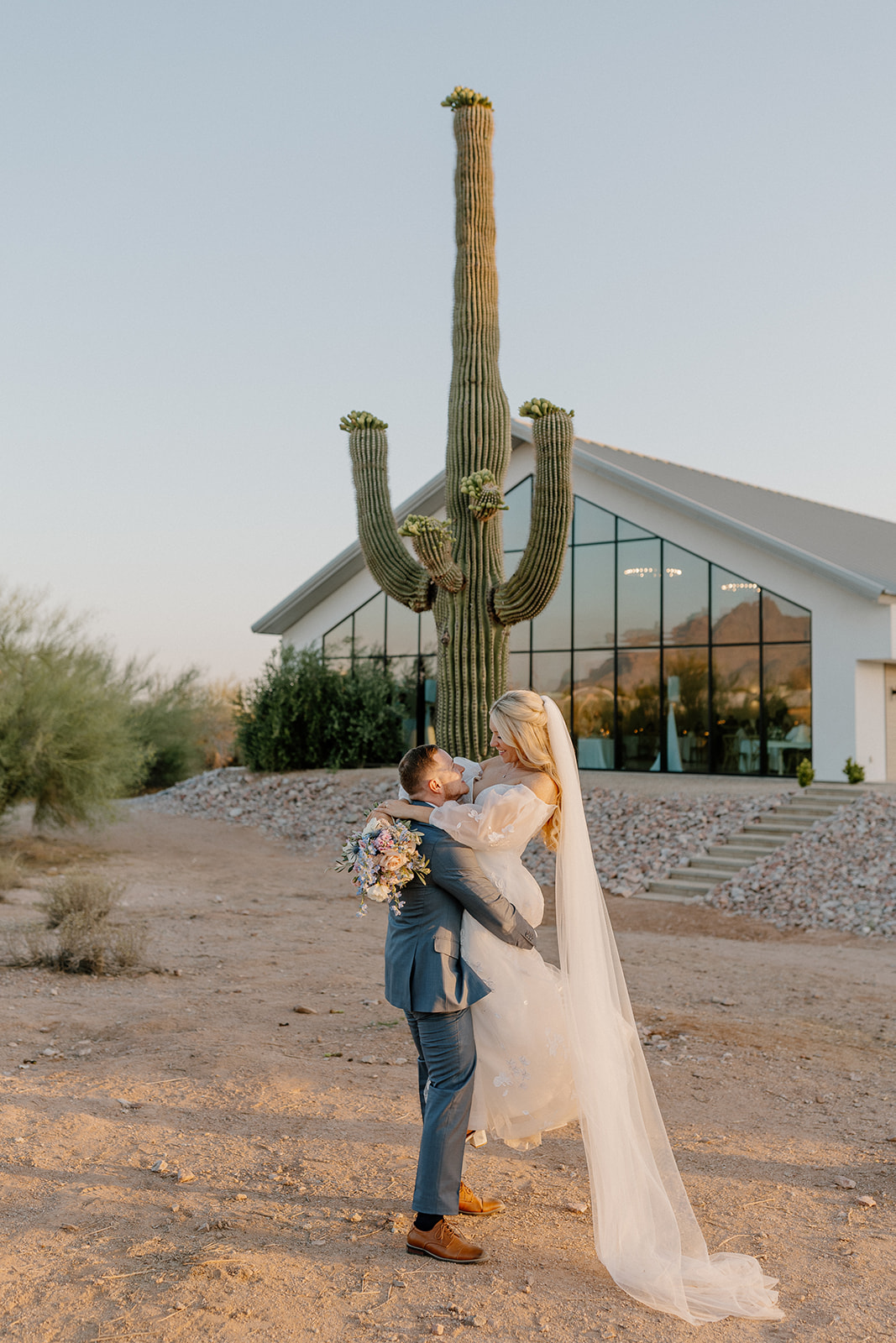 Groom lifts bride in air in front of wedding venue celebrating recent marriage. 