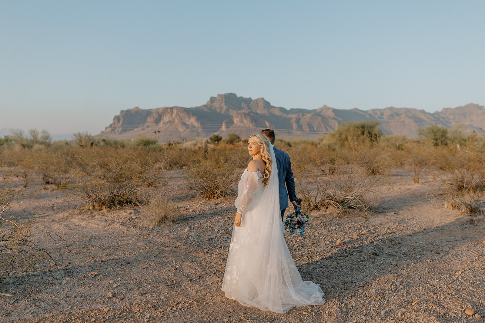 Bride looking over her shoulder while groom leads her into the desert during golden hour portraits. 