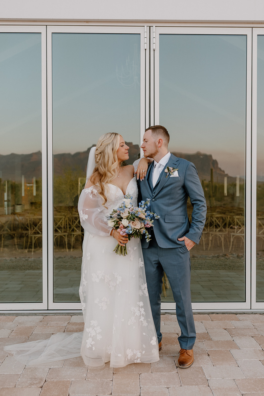 Newly married couple stand looking at one another in front of large windows at wedding venue after wedding ceremony