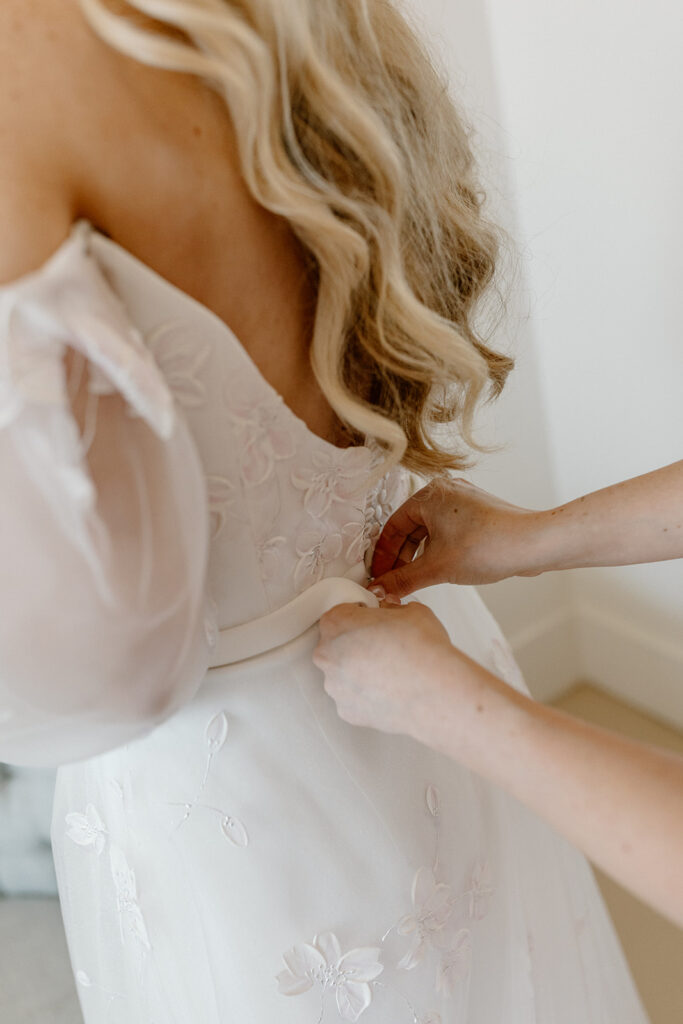 Woman helps bride into her wedding dress by buttoning up her wedding dress. 