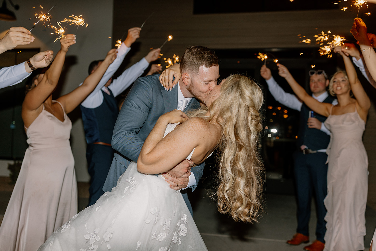 Groom dips the bride to kiss her as guests hold sparklers in the air during their exciting sparkler exit. 