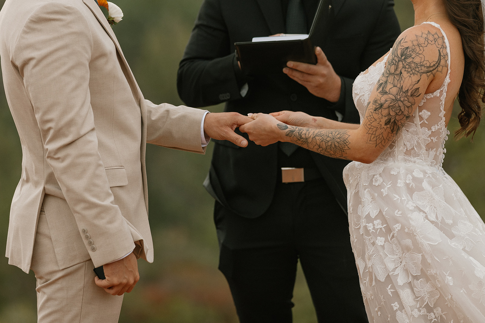Close-up shot of bride and groom holding hands during their wedding ceremony, with the officiant standing behind them and the bride’s floral arm tattoos in full view.