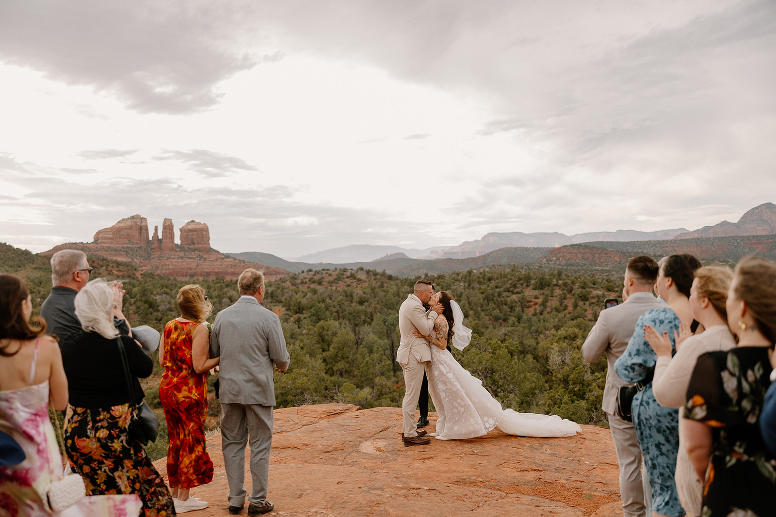 Guests look on and cheer as the couple shares their first kiss on a red rock ledge with Cathedral Rock in the distance, capturing the magic of their Bell Rock Sedona elopement.