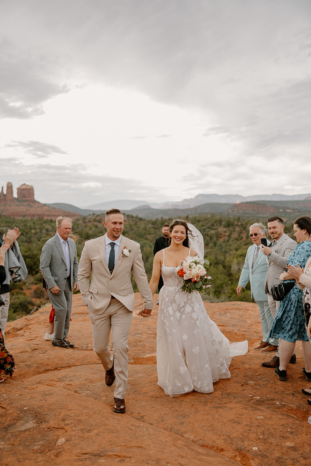 The newlyweds walk joyfully back down the aisle, grinning as friends and family celebrate around them, set against soft sunset skies.