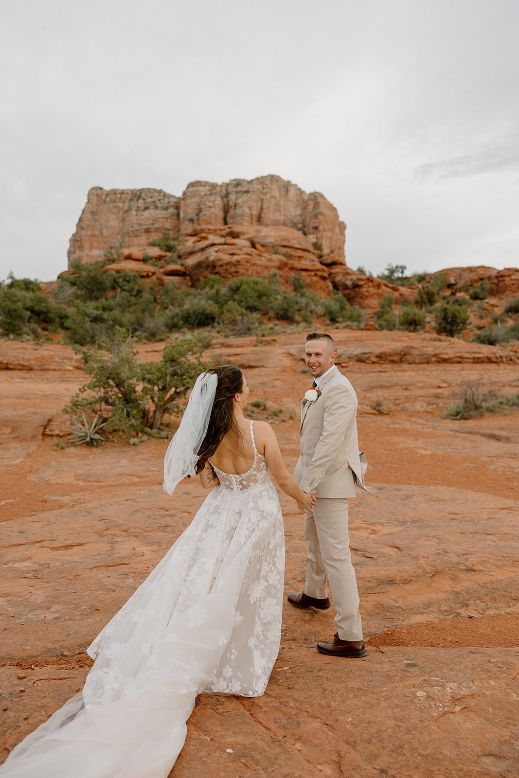 Groom looks back over his shoulder as he holds hands with his bride, leading her across the rust-colored stone with dramatic cliffs behind them.