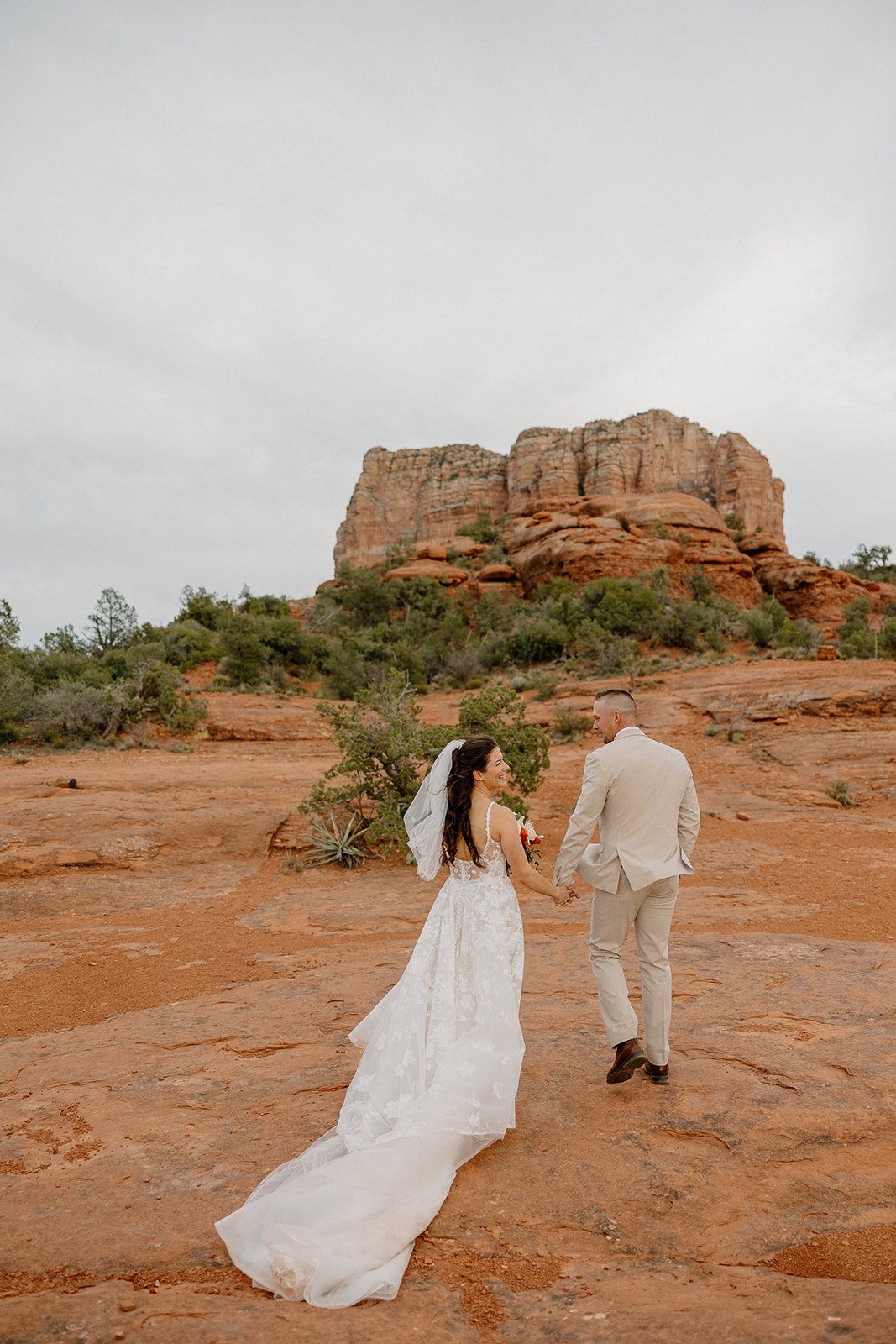 Holding hands and smiling, the couple walks across the sandstone ledge beneath Bell Rock Sedona, surrounded by brush and rust-colored rock.