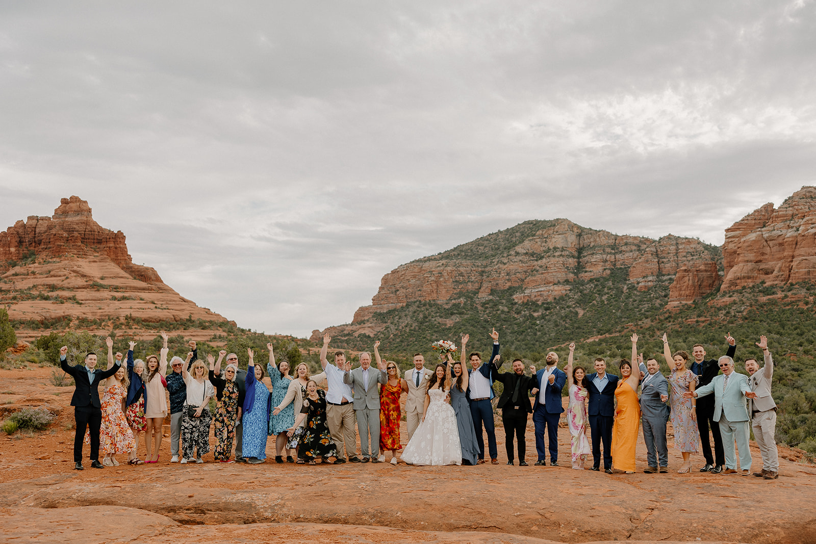 Full wedding group photo on Bell Rock Sedona, guests raising their arms in celebration with red rock buttes and overcast skies creating a cinematic desert backdrop.