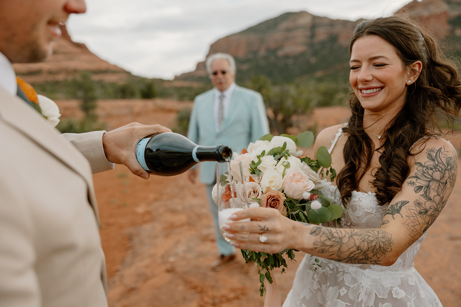 Groom pours champagne into a glass held by the smiling bride, her bouquet tucked under her arm, with Bell Rock Sedona peeking through the background.