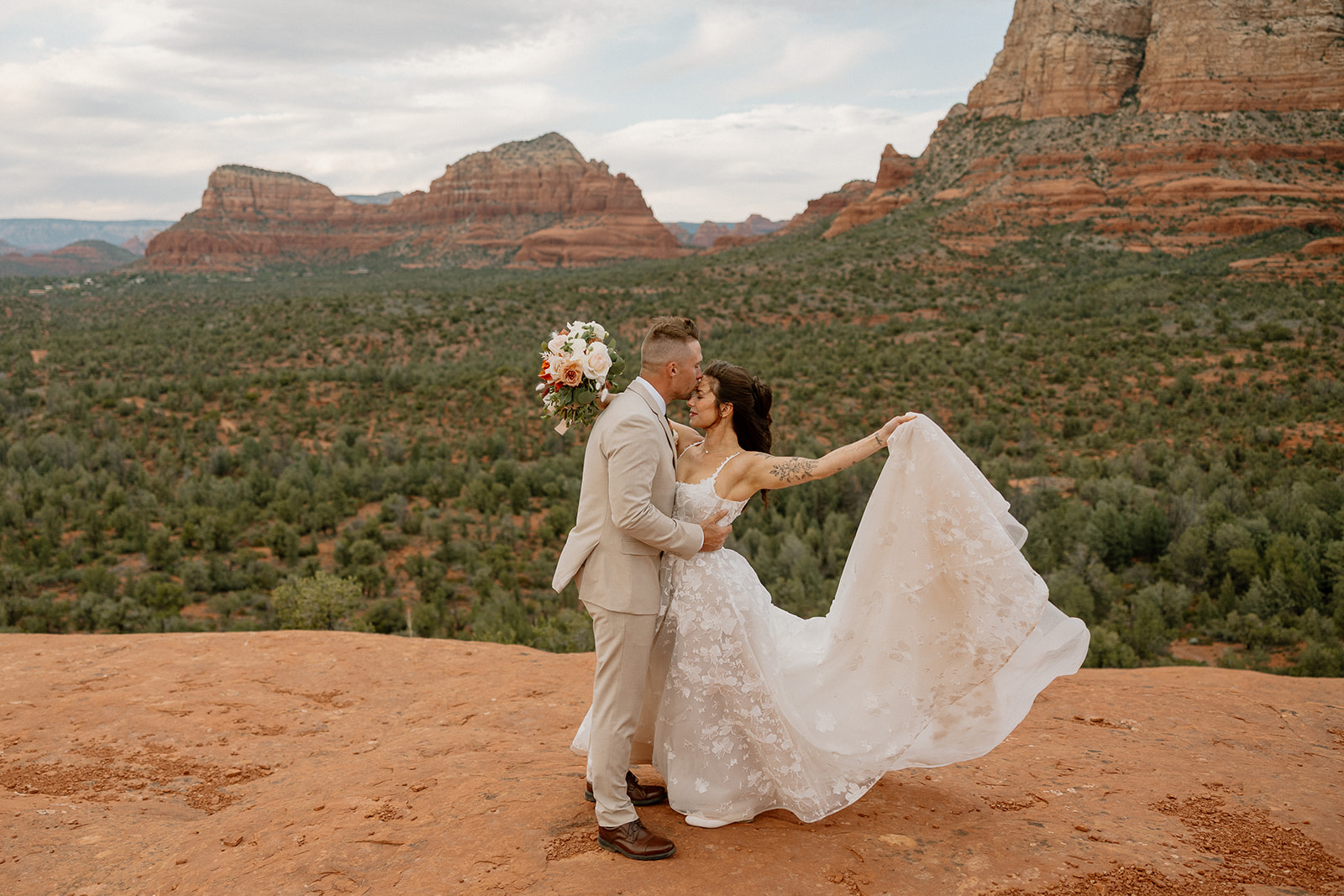 Groom kisses his bride’s forehead while holding a bouquet, her dress billowing as they take in the breathtaking Sedona scenery.