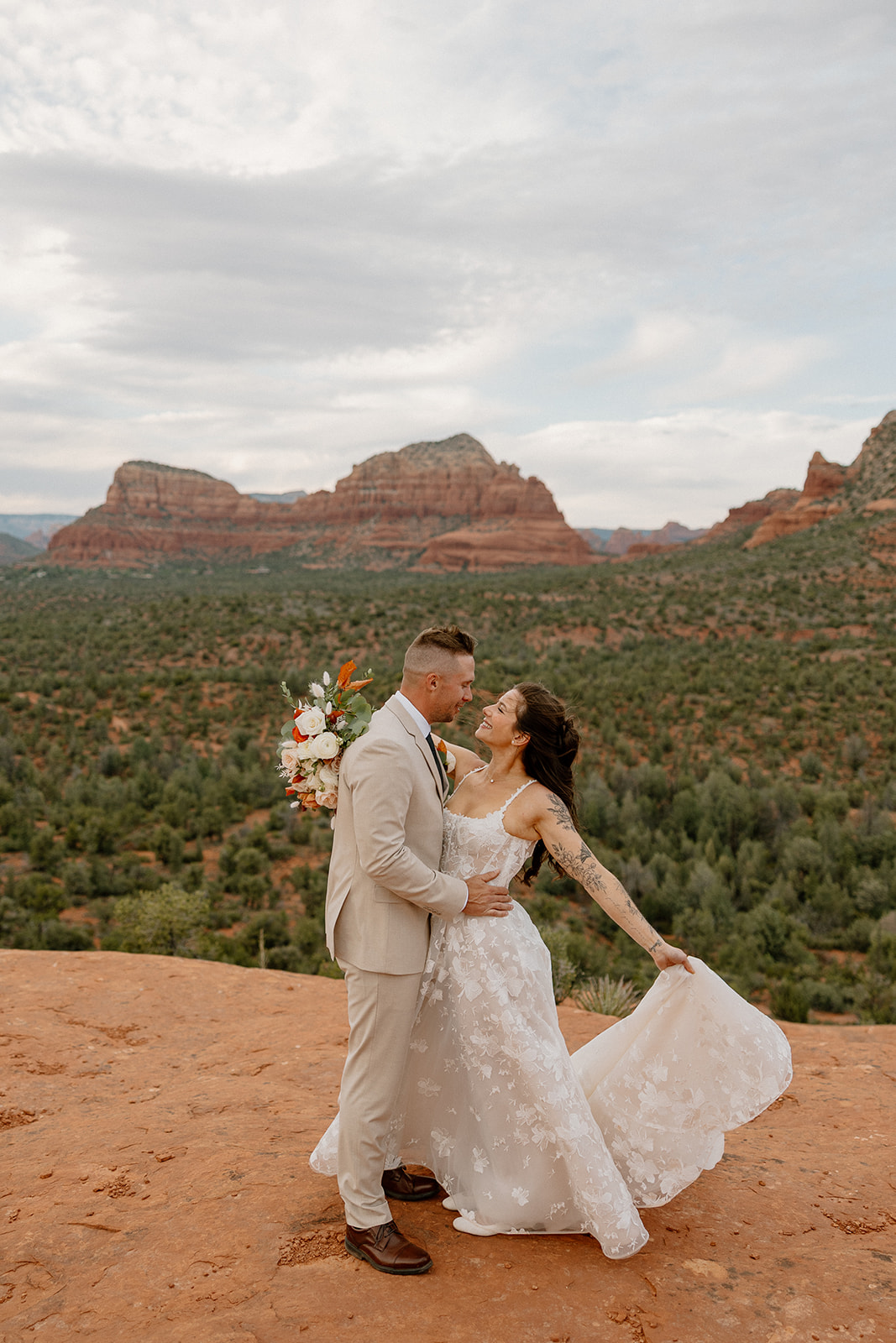 Groom holding bouquet as he gazes at his bride, who twirls slightly with a smile, framed by Bell Rock Sedona’s iconic layered cliffs in the distance.