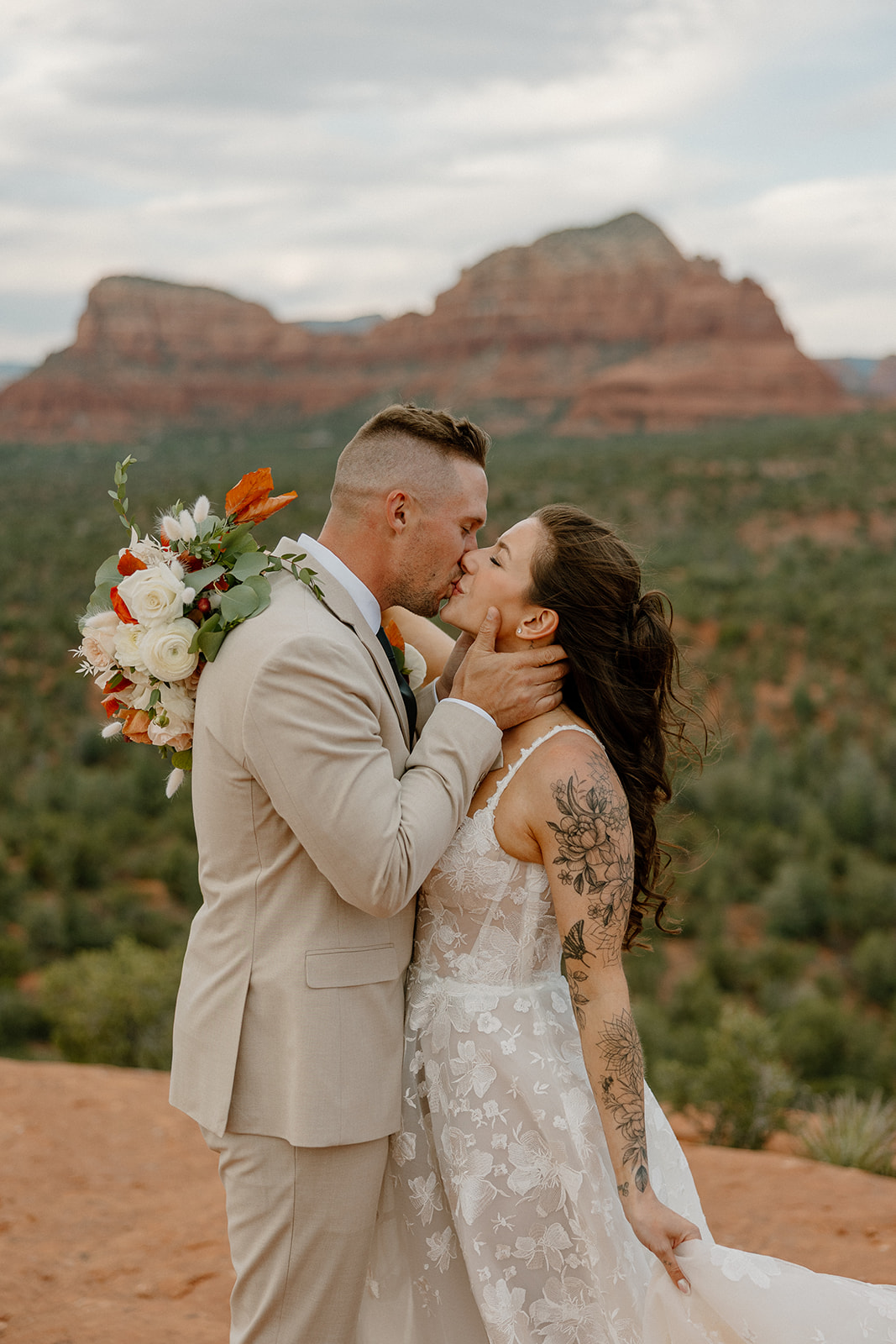 Intimate moment of the couple sharing a kiss, bouquet in hand, with her dress gently flowing and the Sedona desert landscape stretching behind them.