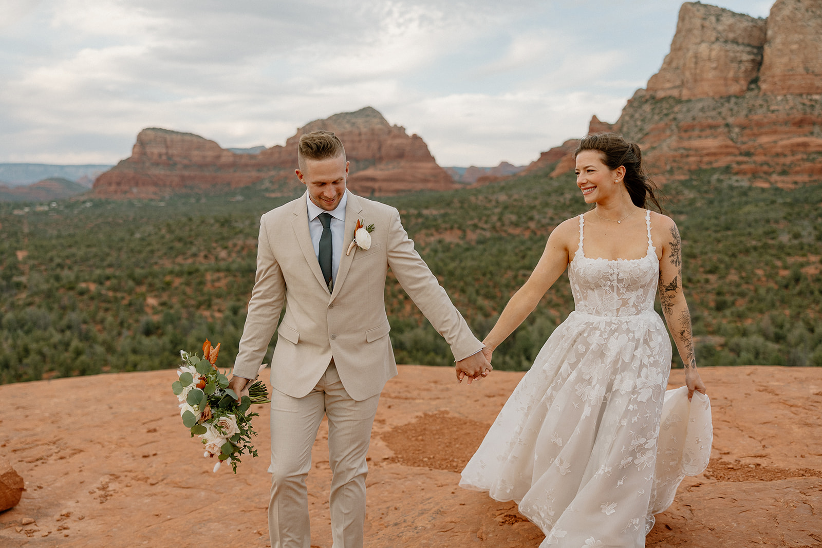 Bride and groom walk hand-in-hand across a cliffside trail, both smiling and windswept, with red rock formations in the background.