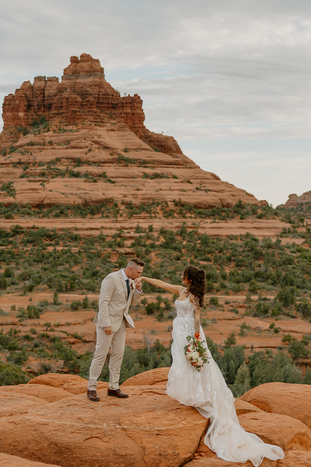 Groom kisses the bride’s hand atop Bell Rock Sedona, her bouquet hanging at her side and layers of sandstone rising behind them in soft morning light.