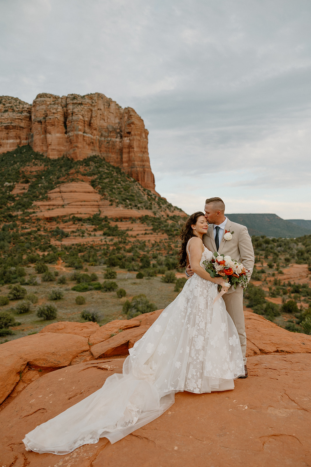The groom kisses his bride’s temple as she smiles and holds a vibrant bouquet, with massive red cliffs towering behind them under a moody sky.