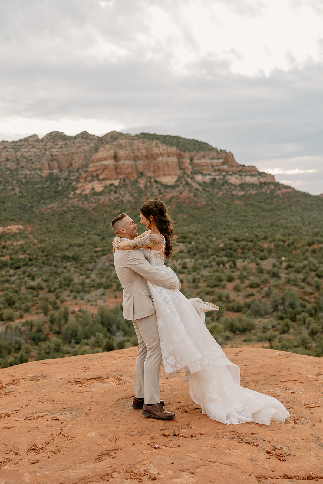 Groom lifts his bride in a playful spin on the cliffs of Bell Rock Sedona, the train of her dress billowing out as the red mountains stretch beyond.