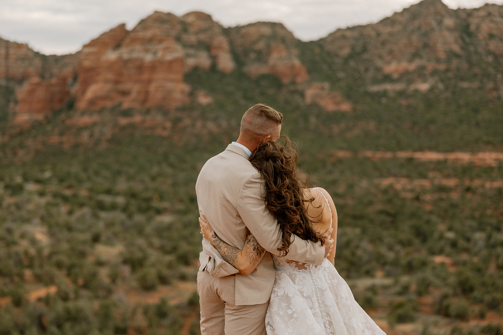 Couple shares a quiet moment with arms wrapped around each other, gazing out over the green desert valley and distant red cliffs of Sedona.
