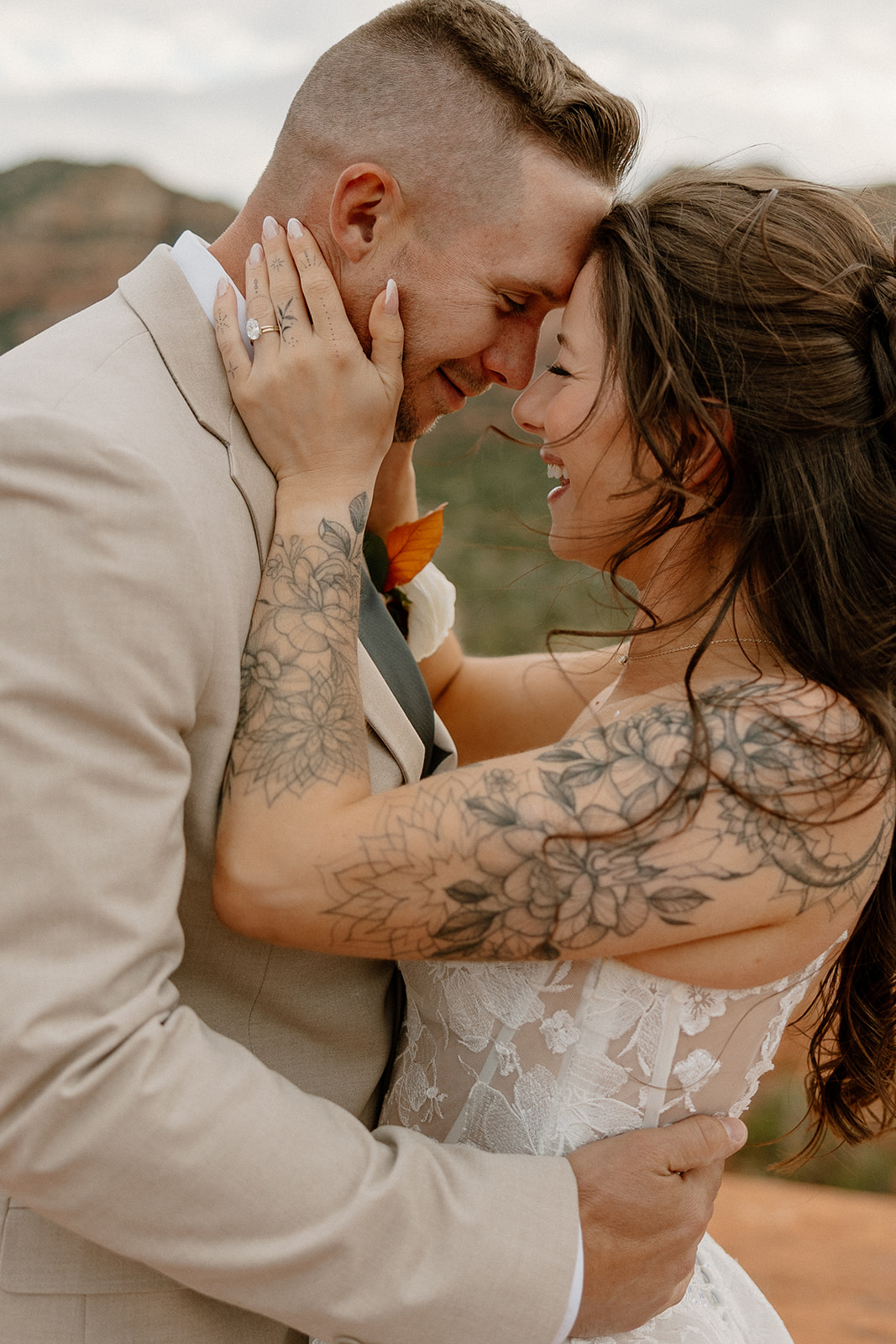 Close-up of a joyful bride and groom sharing a forehead-to-forehead moment, smiling with eyes closed as the bride cradles the groom’s face; her floral tattoos and lace gown are highlighted against a blurred red rock background.