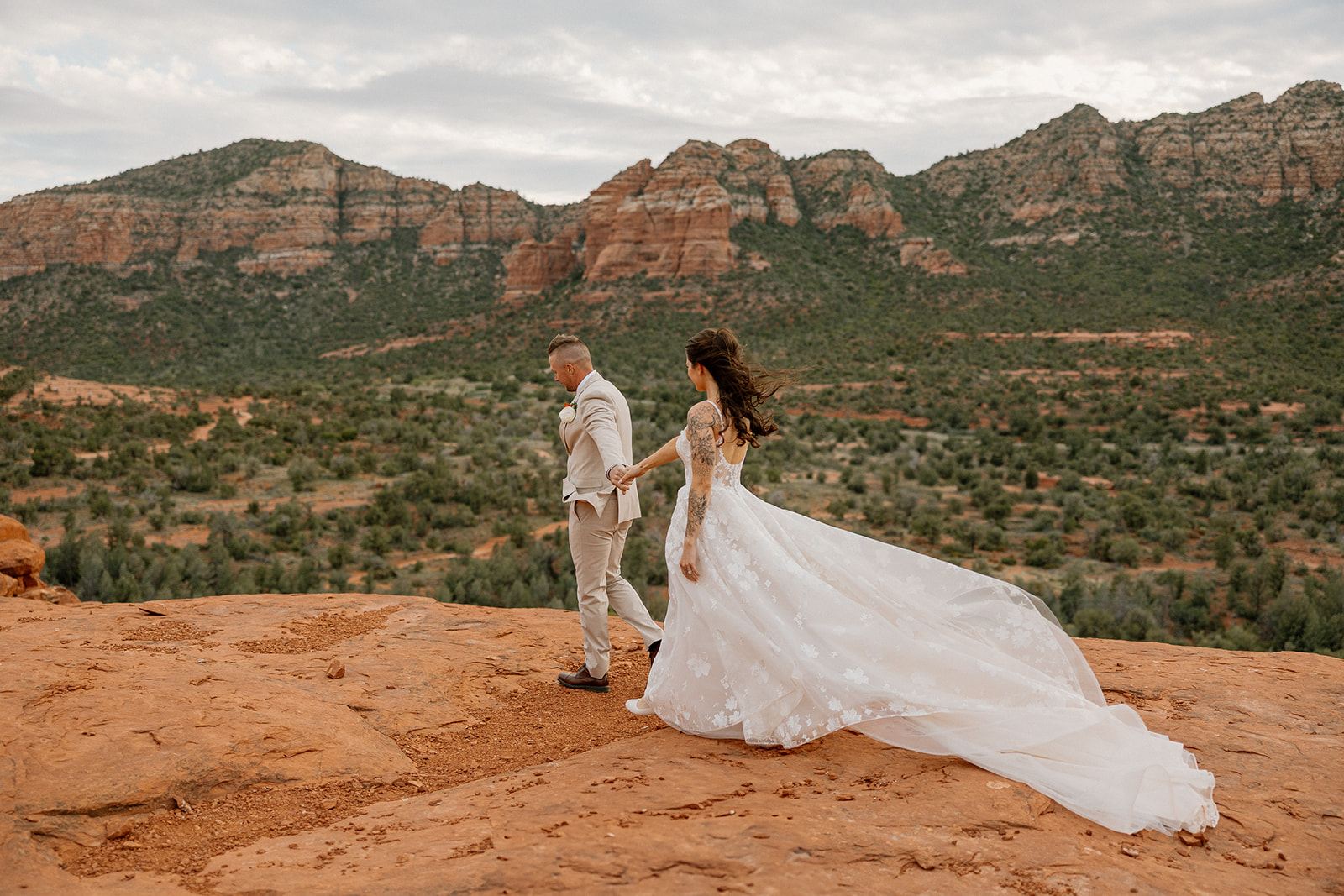 Groom leads his bride across a sweeping red rock cliff, her floral lace train flowing dramatically behind her as they overlook Sedona’s vast desert terrain.