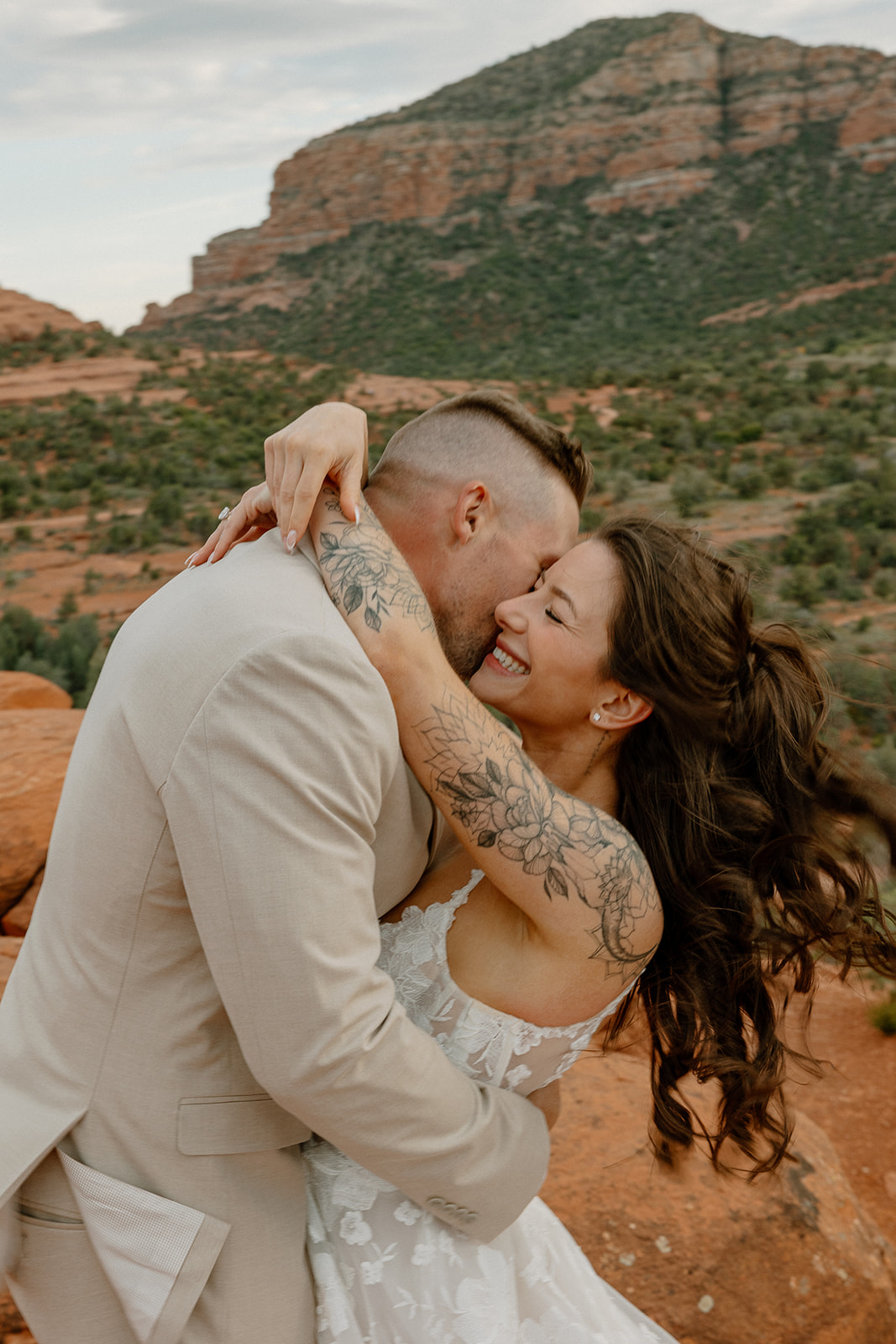 The bride beams as she hugs her groom tight, her tattooed arms wrapped around his shoulders, caught in a windswept embrace among the red rocks.