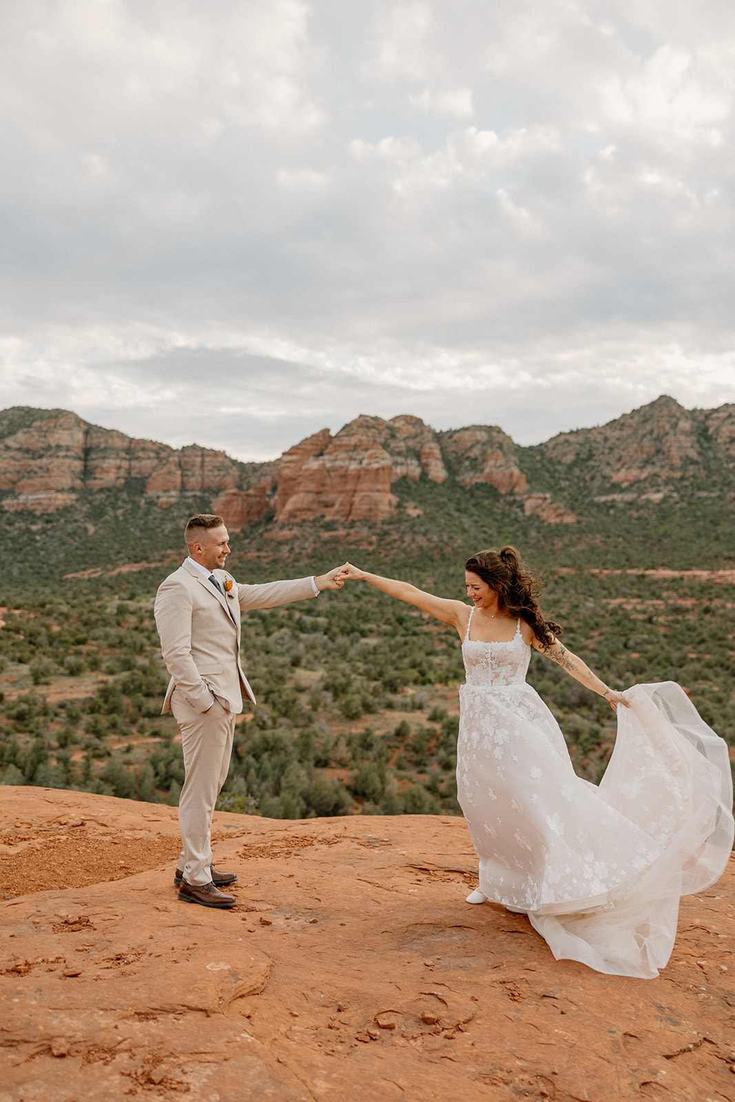 Bride twirls on a red rock ledge in her ethereal dress as the groom watches with a soft smile, both surrounded by the rich green-and-rust hues of the Sedona landscape.